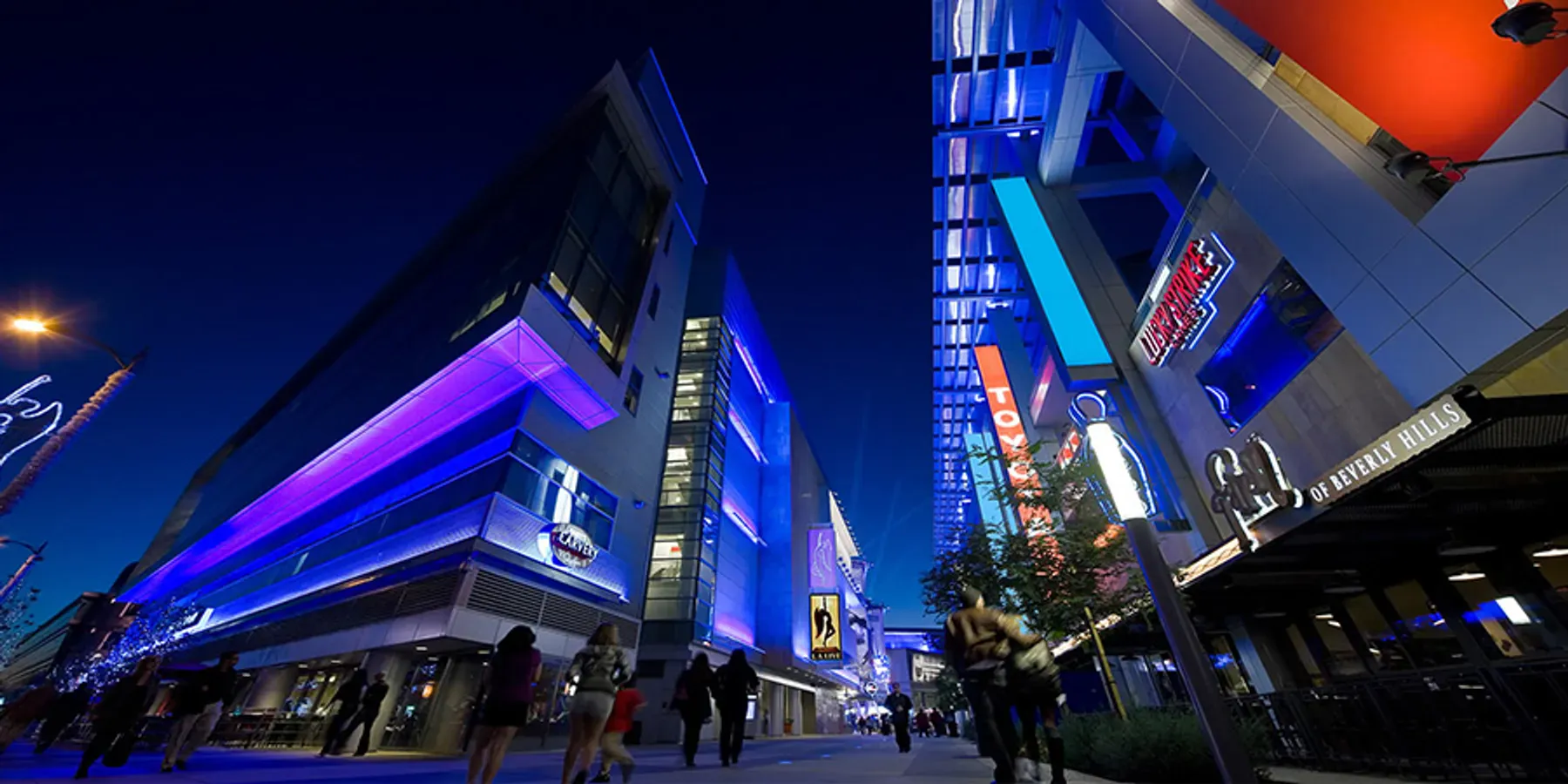 A nighttime view of a multi-story building with blue and purple lighting, surrounded by people on a city street.